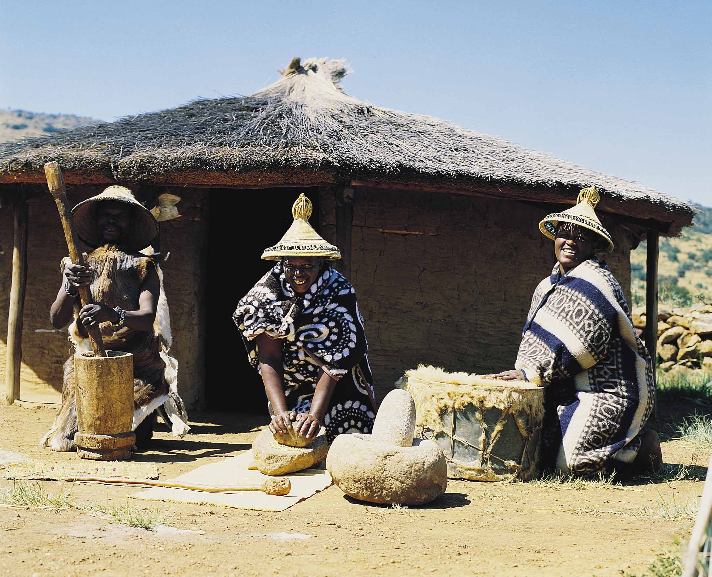 Three women in hats and a basket are outside of a hut.